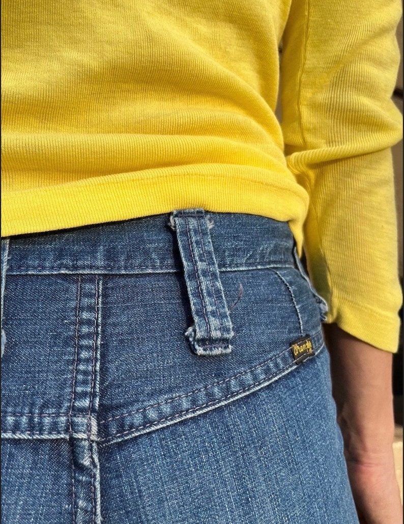 Pu&ograve; includere: Un paio di jeans blu denim con una camicia gialla infilata dentro. I jeans hanno una tasca posteriore con il logo Wrangler.