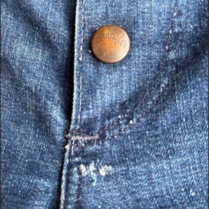 Pu&ograve; includere: Primo piano di un tessuto denim blu con un bottone color rame su cui &egrave; scritto "Wrangler".