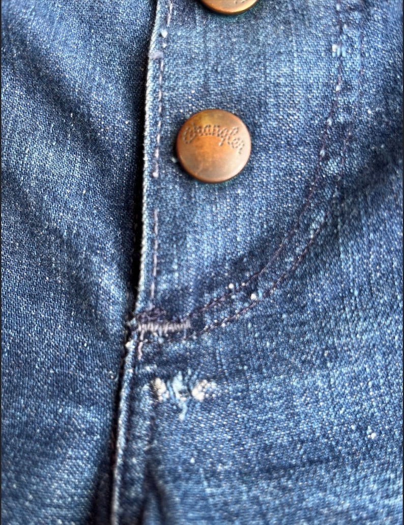 Pu&ograve; includere: Primo piano di un tessuto denim blu con un bottone color rame su cui &egrave; scritto "Wrangler".