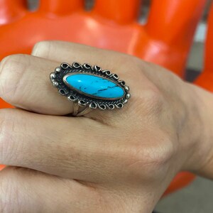 Puede incluir: Un anillo de plata con una piedra ovalada turquesa engastada en un engaste de plata decorativo. El anillo est&aacute; en un dedo.