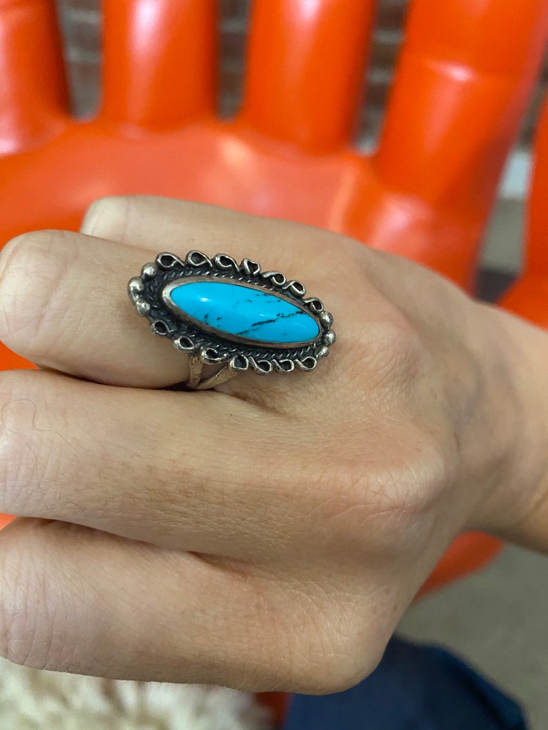 Puede incluir: Un anillo de plata con una piedra ovalada turquesa engastada en un engaste de plata decorativo. El anillo est&aacute; en un dedo.