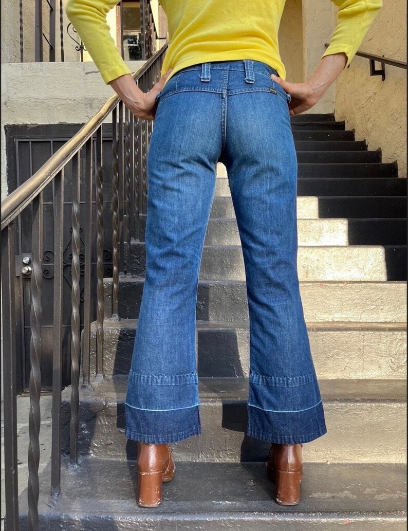 Pu&ograve; includere: Un paio di jeans blu denim a gamba larga con orlo a pie rovesciato. I jeans hanno vita alta e chiusura con bottone.
