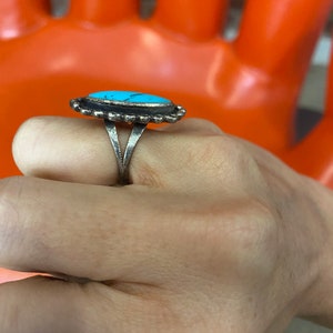 Puede incluir: Un anillo de plata con una piedra turquesa engastada en un engaste de bisel. El anillo tiene una banda decorativa con un borde festoneado.