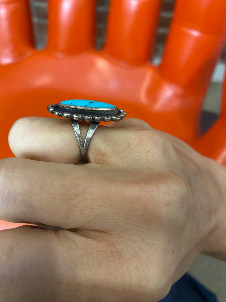 Puede incluir: Un anillo de plata con una piedra turquesa engastada en un engaste de bisel. El anillo tiene una banda decorativa con un borde festoneado.