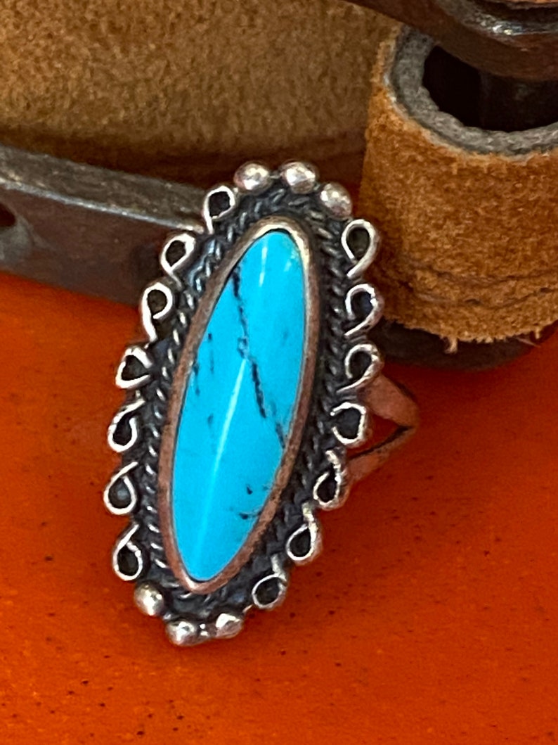 Puede incluir: Un anillo de plata con una piedra turquesa engastada en forma ovalada. El anillo tiene un borde decorativo alrededor de la piedra.