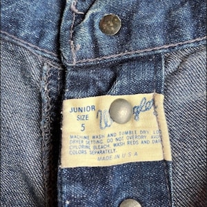 Pu&ograve; includere: Un primo piano di una cintura in denim blu con un'etichetta bianca che recita "JUNIOR SIZE 5" e istruzioni per il lavaggio e l'asciugatura del capo. L'etichetta &egrave; fissata con due bottoni argentati.