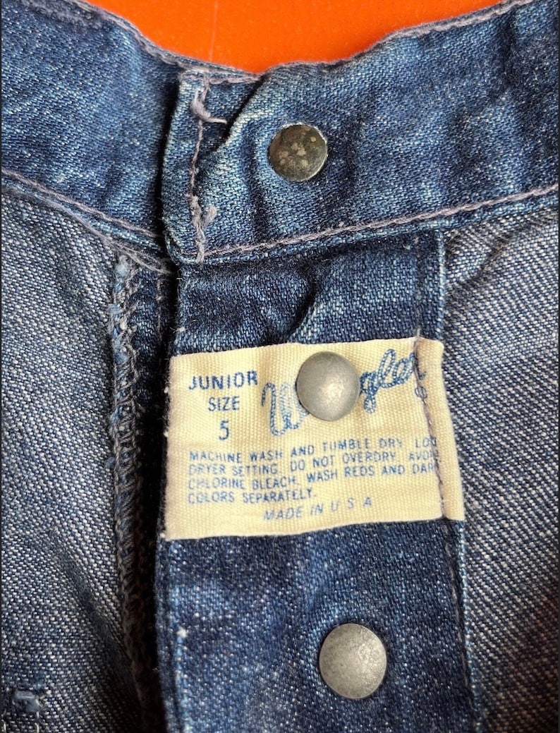 Pu&ograve; includere: Un primo piano di una cintura in denim blu con un'etichetta bianca che recita "JUNIOR SIZE 5" e istruzioni per il lavaggio e l'asciugatura del capo. L'etichetta &egrave; fissata con due bottoni argentati.