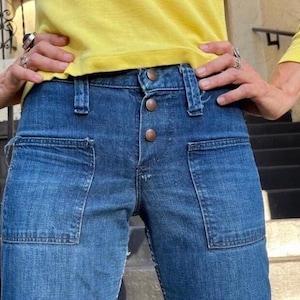 Pu&ograve; includere: Un paio di jeans blu denim con grandi tasche anteriori e chiusura con bottoni. I jeans sono indossati da una persona con una camicia gialla.