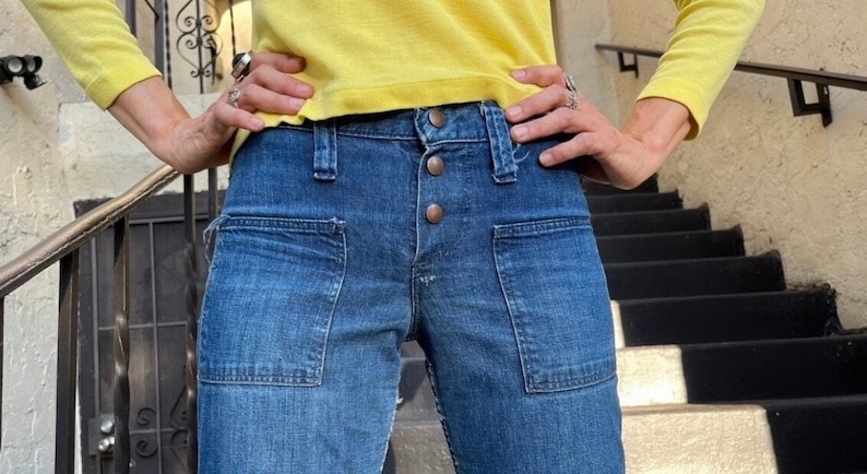 Pu&ograve; includere: Un paio di jeans blu denim con grandi tasche anteriori e chiusura con bottoni. I jeans sono indossati da una persona con una camicia gialla.