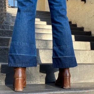 Pu&ograve; includere: Un paio di jeans a zampa d'elefante in denim blu con un dettaglio di cucitura blu chiaro. I jeans sono indossati con stivali marroni con tacco.