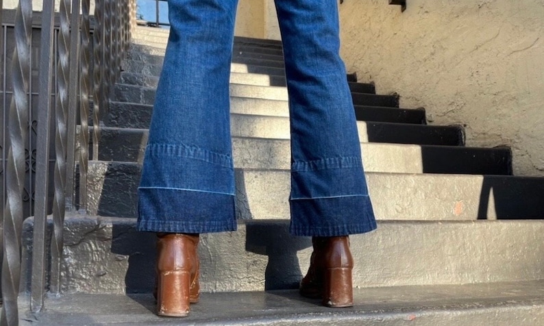 Pu&ograve; includere: Un paio di jeans a zampa d'elefante in denim blu con un dettaglio di cucitura blu chiaro. I jeans sono indossati con stivali marroni con tacco.
