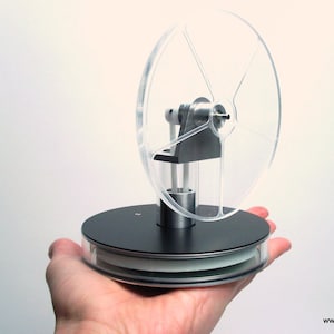 Puede incluir: Un modelo transparente de motor Stirling con un volante transparente y una base negra. El motor tiene componentes metálicos plateados y se sostiene en una mano sobre un fondo blanco. La dirección del sitio web "www.exergia.de" es visible.
