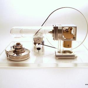 May include: A clear glass tube with a silver and gold engine attached to a clear acrylic base. The engine has a clear plastic cover and a silver flywheel. The base has a silver and gold mechanism on the left side.