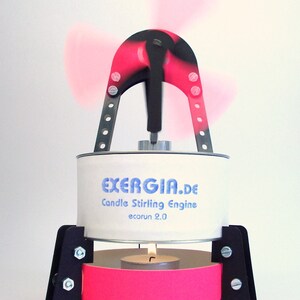Könnte beinhalten: Eine weiße Metalldose mit dem Text "EXERGIA.de Candle Stirling Engine ecorun 2.0" darauf gedruckt. Die Dose ist an einem schwarzen Metallrahmen befestigt, auf dem sich oben ein roter und schwarzer Propeller dreht. Am unteren Ende des Rahmens ist eine kleine Teelichtkerze zu sehen.