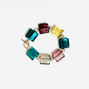 Pode incluir: Um bracelete feito de contas quadradas de vidro coloridas. As contas são em tons de azul-esverdeado, amarelo, rosa e bordô. O bracelete tem um fecho dourado.