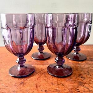 May include: A set of four purple glass goblets featuring a vertical ribbed pattern on the bowl, a faceted stem, and a circular base. The glassware has a deep violet hue and a translucent finish, with two glasses positioned in the front and two behind.