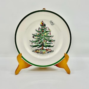 May include: A white ceramic plate with a green border and a Christmas tree design. The tree is decorated with ornaments and sits above wrapped presents. The plate is displayed on a wooden stand.