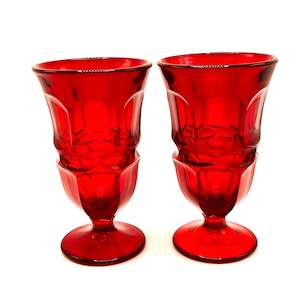 May include: Two ruby red vintage drinking glasses. Each glass has a pedestal base, a faceted body, and a flared rim. The glasses are made of a transparent material and are set against a white background. These glasses are perfect for serving beverages.
