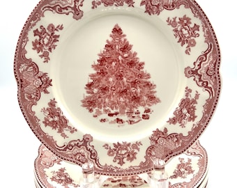 May include: A stack of cream-colored ceramic plates with a scalloped edge. The center of the top plate features a detailed red illustration of a decorated Christmas tree with gifts underneath. The rim is decorated with a red floral pattern.