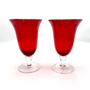 May include: Two red-colored stemmed glasses with a clear, rounded base. The glasses have a tulip-shaped bowl and a clear stem. The glasses are made of glass and are set against a white background.