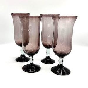 May include: A set of four translucent purple glass goblets with flared rims, clear glass stems, and wide, dark purple circular bases. Each goblet features a smooth, curved bowl shape and a distinct transition between the colored glass and stem.