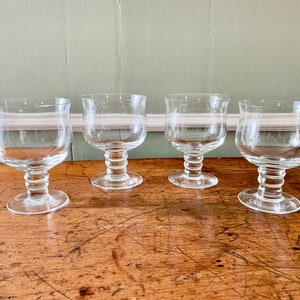 May include: Four clear glass drinking glasses with a wide bowl and a short stem. The glasses are sitting on a wooden surface.