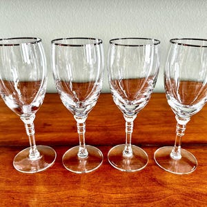May include: Four clear glass wine glasses with a silver rim. The glasses are arranged in a row on a wooden surface.