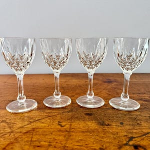May include: Four clear crystal wine glasses with intricate etched patterns. The glasses have a classic design with a stemmed base and a rounded bowl. They are arranged on a wooden surface.