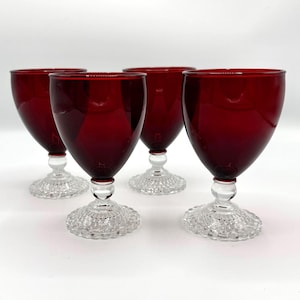 May include: Four ruby red wine glasses with clear, textured bases. The glasses have a classic goblet shape, with a deep red bowl and a clear stem. The bases are intricately patterned, adding a touch of elegance to the set.