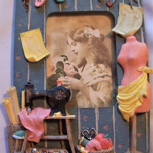 Może przedstawiać: Niebieska i biała ramka do zdjęć z motywem szycia. Ramka zawiera maszynę do szycia, manekin i zdjęcie młodej kobiety trzymającej kwiat. Ramka jest ozdobiona guzikami, szpilkami i szpilką.