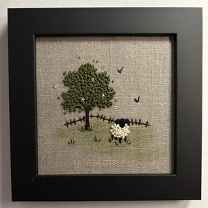 May include: Embroidered artwork featuring a black and white sheep standing in a field with a green tree and two birds flying overhead. The artwork is framed in black.