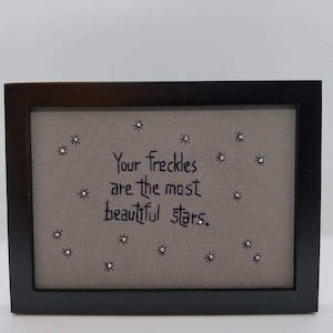 May include: A framed embroidery with the text "Your freckles are the most beautiful stars."  The embroidery is on a light gray fabric with silver glitter stars.