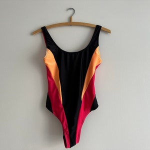 May include: A black swimsuit with orange and red colour blocking. The swimsuit has a scoop neck and thin straps.