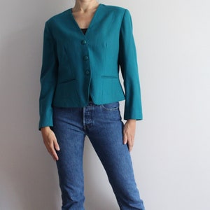 May include: A teal blazer with a v-neck and four buttons. The blazer is fitted and has a cropped length. The model is wearing blue jeans.