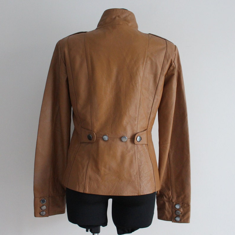 May include: A brown leather jacket with a stand-up collar and a button closure. The jacket has a fitted silhouette and features a row of buttons on the back.