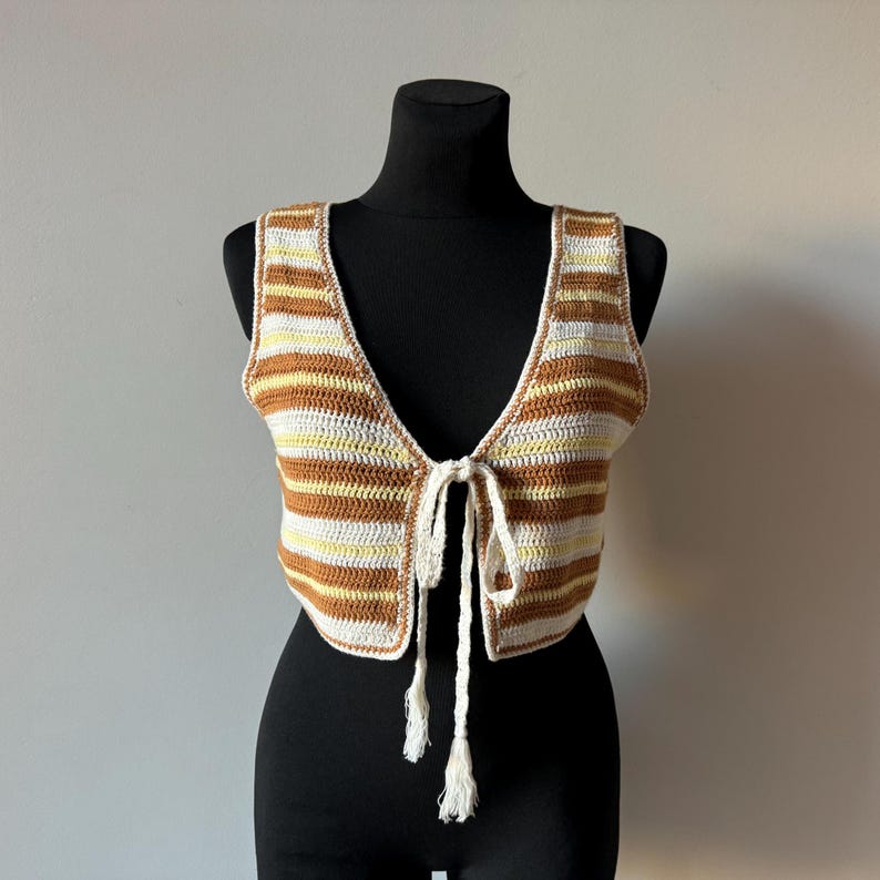 Puede incluir: Un chaleco de punto de crochet a rayas marrones y blancas con un frente de corbata y borlas. El chaleco es sin mangas y tiene un escote en V profundo.
