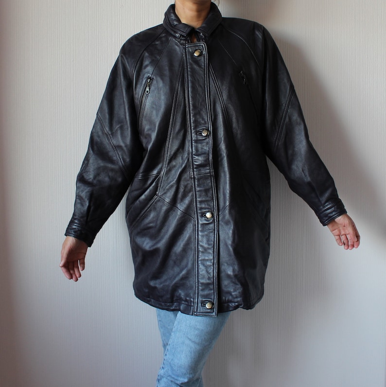 May include: A black leather jacket with a button-up front and multiple pockets. The jacket has a long, oversized fit.