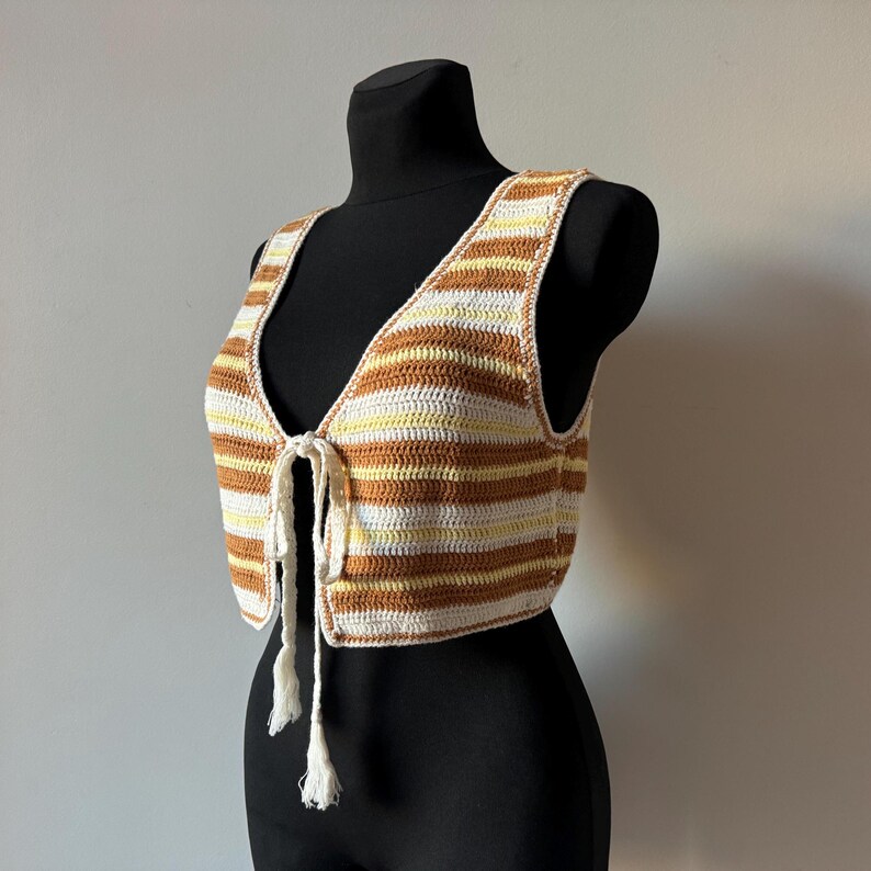 Puede incluir: Un chaleco de crochet a rayas marrones y blancas con un frente de corbata. El chaleco es sin mangas y tiene una longitud recortada.