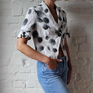 May include: A white blazer with a black polka dot pattern. The blazer has short sleeves, a notched collar, and black buttons. The person is wearing blue jeans.