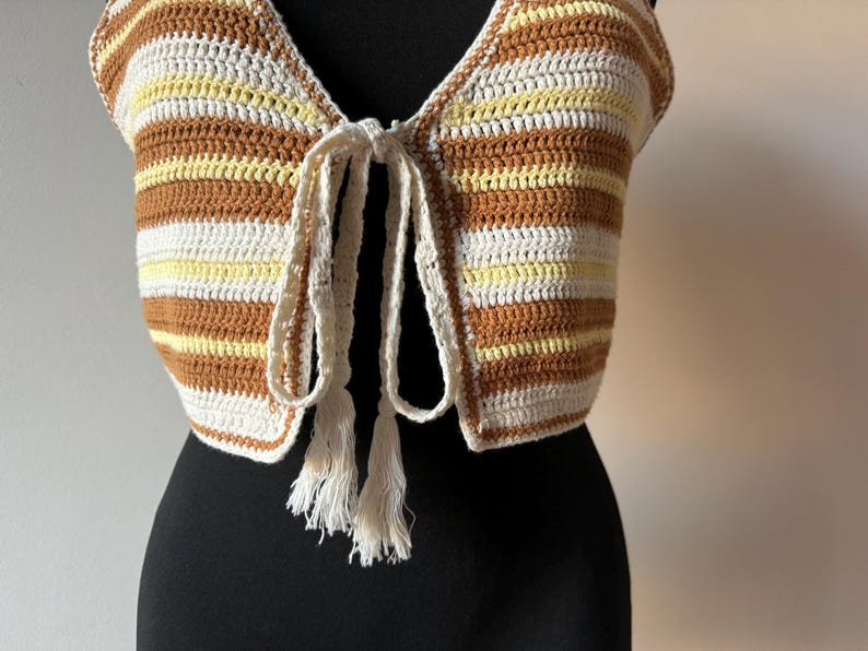 Puede incluir: Un top de crochet con un frente de corbata y borlas. El top est&aacute; rayado en marr&oacute;n, blanco y amarillo.