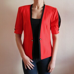Könnte beinhalten: Ein roter Blazer mit schwarzen Besätzen und kurzen Ärmeln. Der Blazer hat einen Knopfverschluss und goldene Knöpfe.