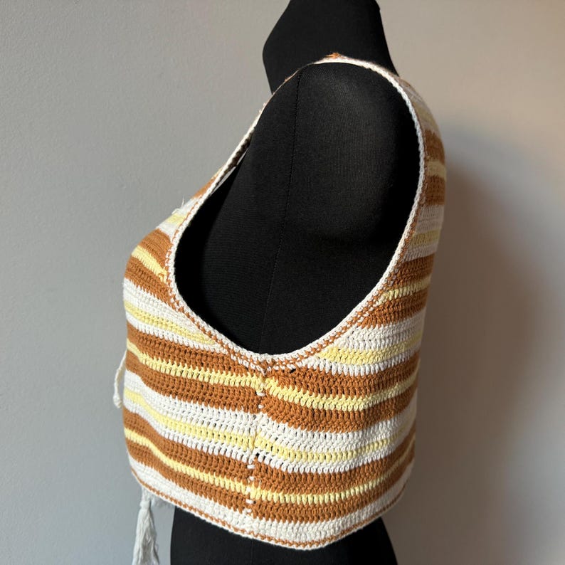 Puede incluir: Un top corto de crochet con rayas blancas y marrones. El top tiene cuello en V y es sin mangas.