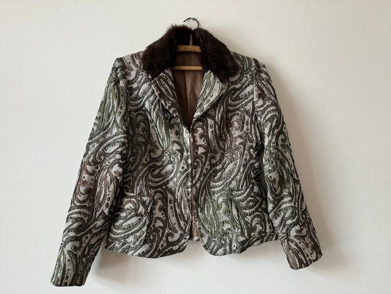K&ouml;nnte beinhalten: Eine braune und gr&uuml;ne gemusterte Jacke mit einem braunen Pelzkragen. Die Jacke hat einen Rei&szlig;verschluss und lange &Auml;rmel.