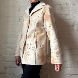 May include: A beige faux suede jacket with a cream-coloured faux fur collar and lining. The jacket features a floral pattern in shades of brown and tan. It has a button closure and a pocket on the front.