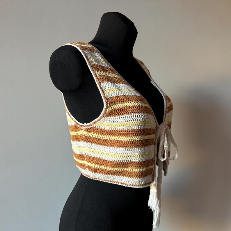 Puede incluir: Un chaleco de punto de crochet a rayas marrones y blancas con cierre de corbata. El chaleco es sin mangas y tiene una longitud recortada.