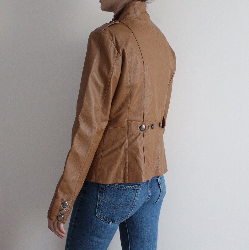 May include: A brown leather jacket with a stand-up collar and button closures. The jacket has a fitted silhouette and a slightly cropped length.