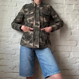 Puede incluir: Una chaqueta utilitaria con estampado de camuflaje que presenta cuello, mangas largas, dos bolsillos en el pecho con solapas y dos bolsillos delanteros inferiores con solapas. La chaqueta incluye un cordón en la cintura y shorts de mezclilla.