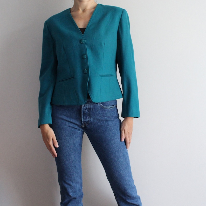 May include: A teal blazer with a v-neck and four buttons. The blazer is fitted and has a cropped length. The model is wearing blue jeans.