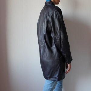 May include: A long, dark brown leather jacket with a collar and button closure. The jacket has a relaxed fit and a slightly oversized look.