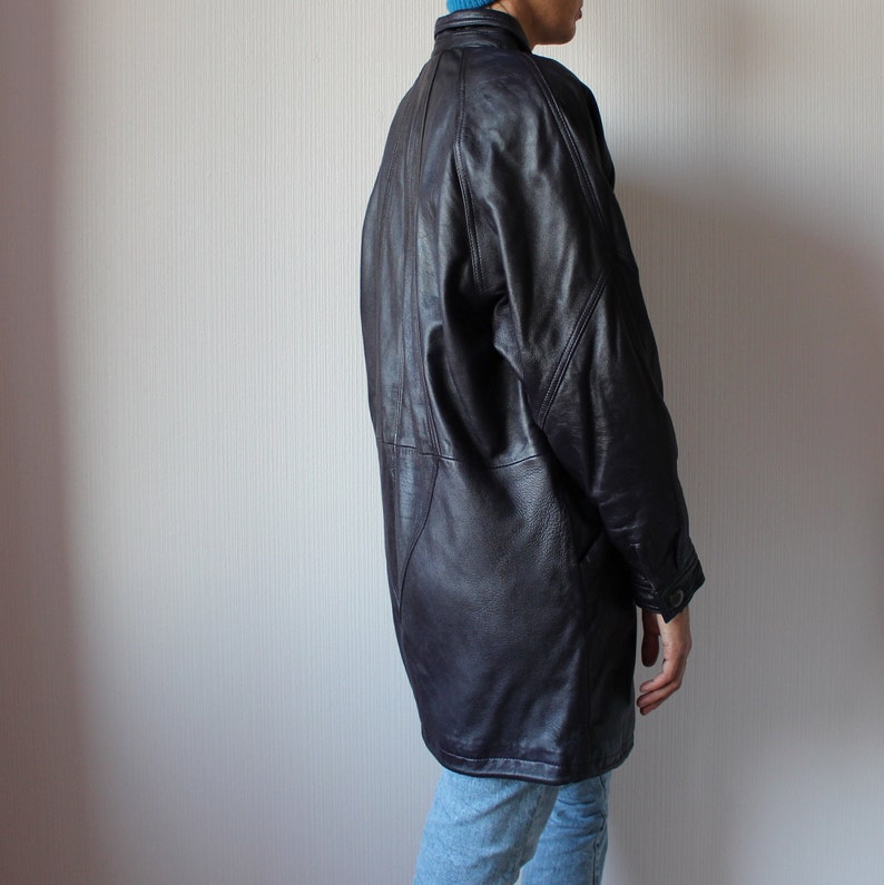May include: A long, dark brown leather jacket with a collar and button closure. The jacket has a relaxed fit and a slightly oversized look.